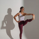 Woman in white tank top and maroon leggings performing a yoga pose against a plain background